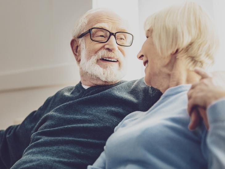 Een senior koppel zit op de bank en kijkt elkaar blij aan, de man heeft zijn arm om de schouders van zijn vrouw geslagen.